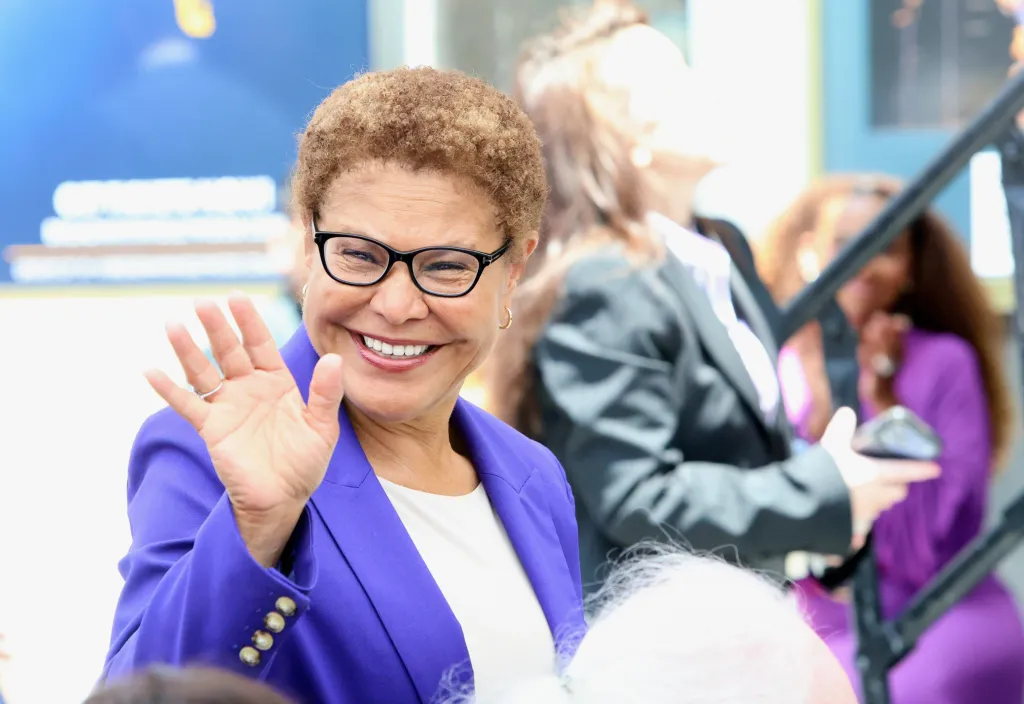 Karen Bass, wearing glasses and a purple jacket, smiles and waves to the camera.