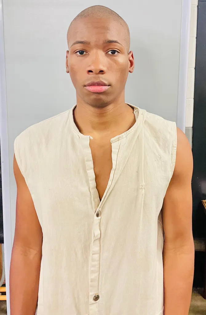 A man with a shaved head and a light-colored sleeveless shirt looks directly at the viewer.