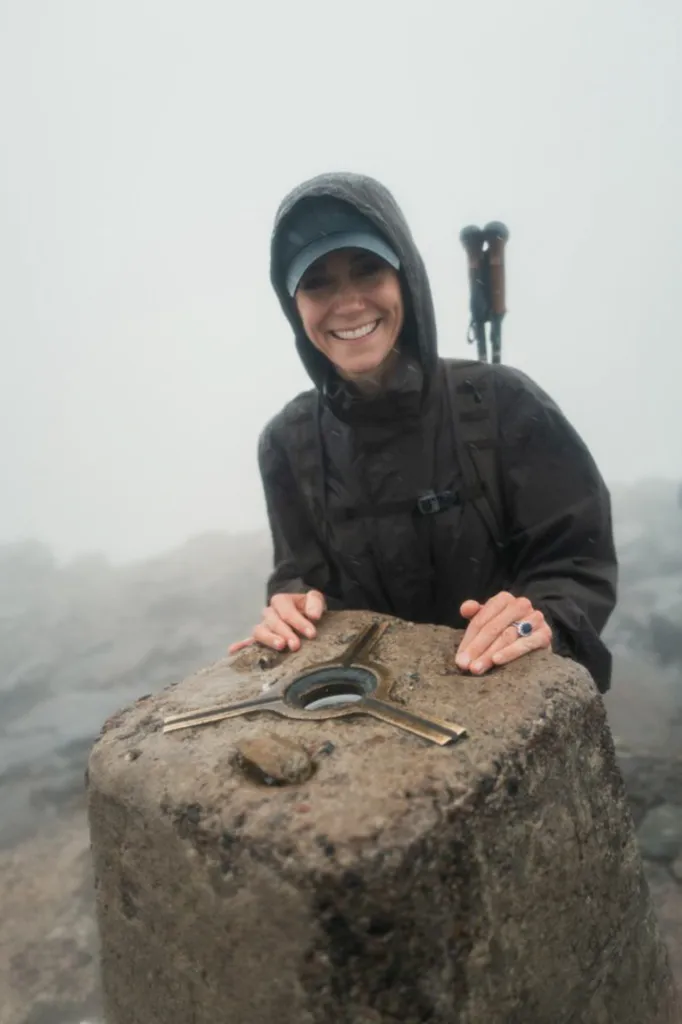Kate Middleton smiling, wearing a rain jacket and cap, with her hands on a stone marker in foggy weather.