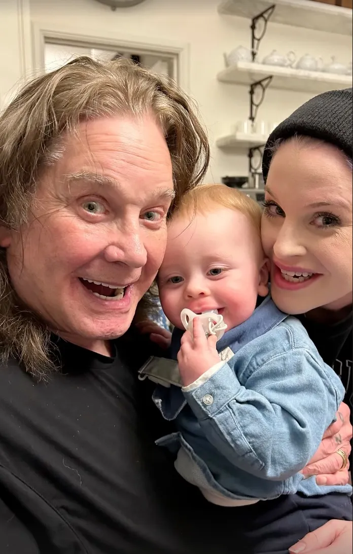 Ozzy Osbourne smiling while holding his grandson, with Kelly Osbourne smiling next to them.