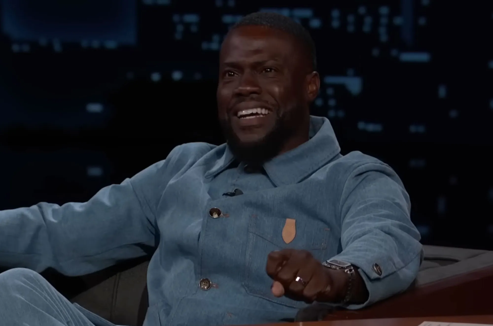 Kevin Hart smiling and laughing while sitting in a blue denim outfit.