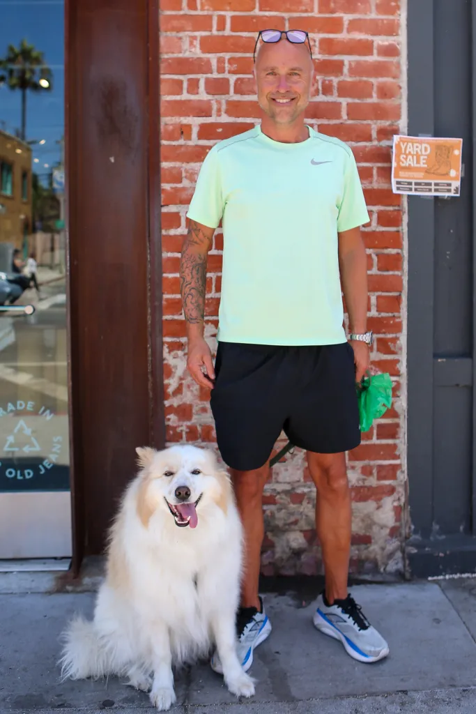Khris Cuyver stands next to a fluffy white dog against a brick wall.