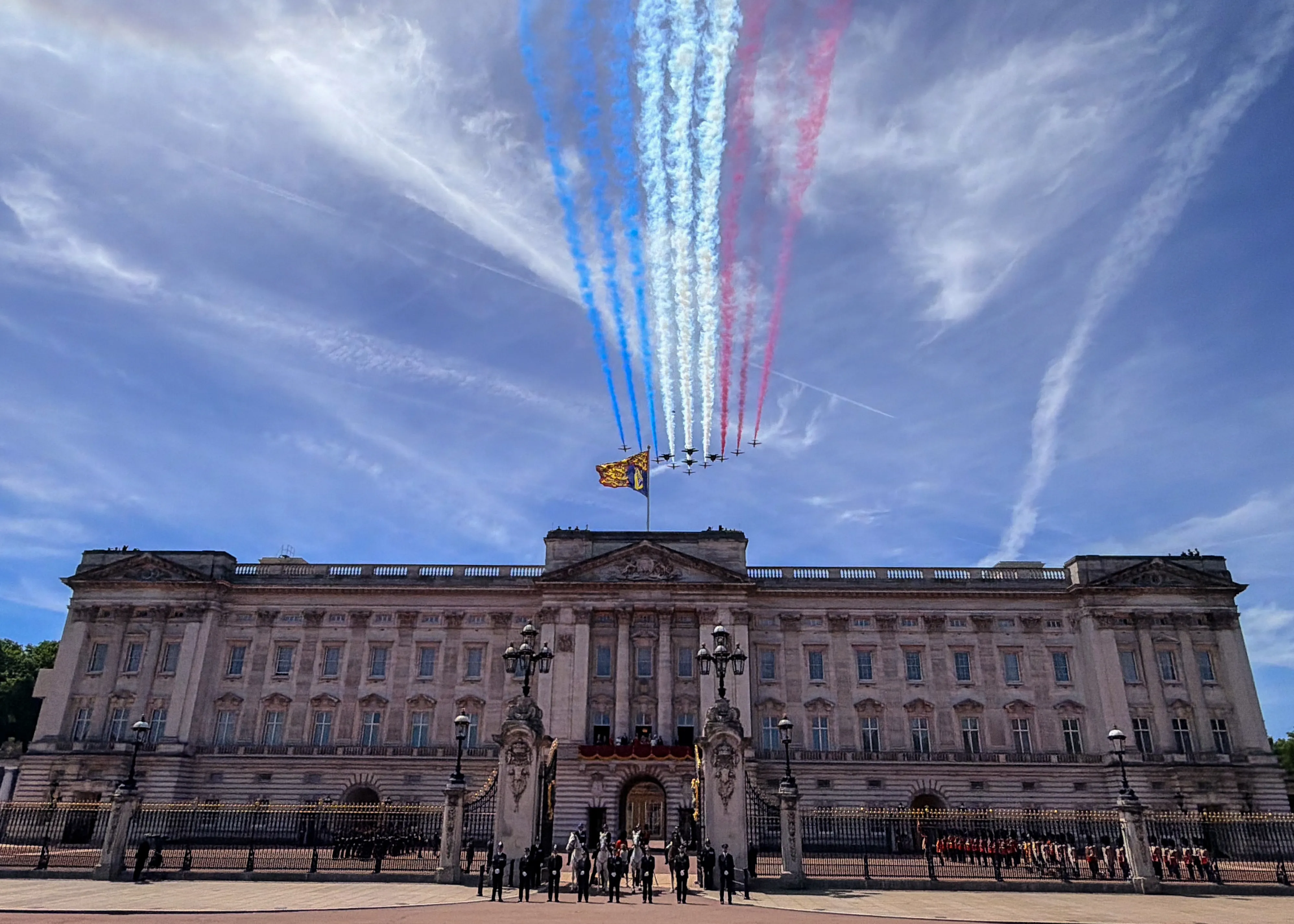 Buckingham Palace with a Royal Standard flag flying and a military aircraft flypast leaving red, white, and blue trails in the sky for Trooping the Colour.