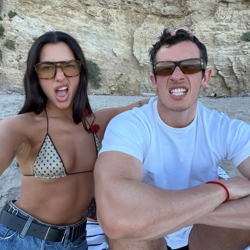 Dua Lipa and Callum Turner on a beach, both wearing sunglasses and making funny faces at the camera.