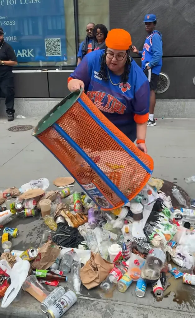 A Knicks fan wearing an orange hat and a blue and orange jersey tips over a blue and orange trash can, spilling its contents onto the street.