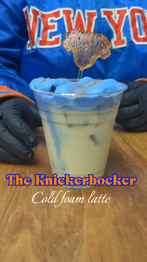 The Knickerbocker cold foam latte, topped with a New York Knicks logo sugar cookie, held by a person in a Knicks jacket.