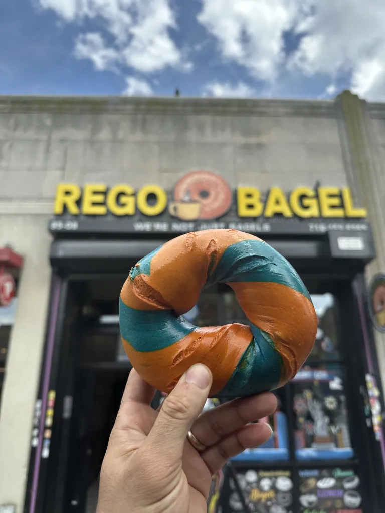Hand holding an orange and blue twist bagel in front of a bagel shop.
