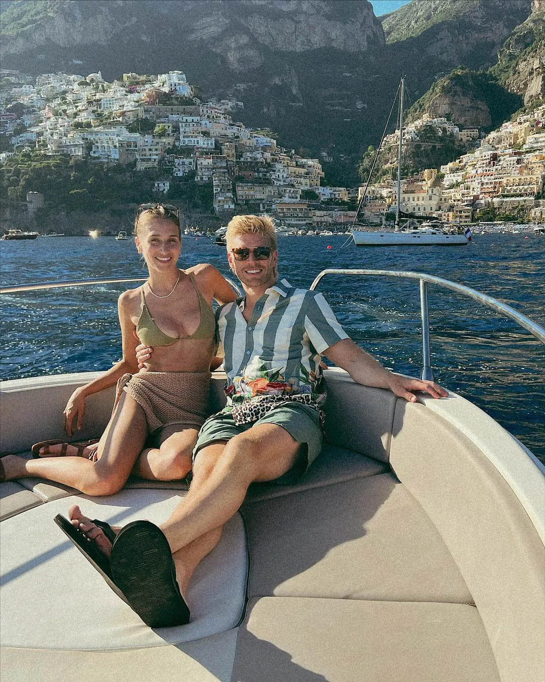 Kyle Cooke and Amanda Batula sit on a boat with a coastal town in the background.