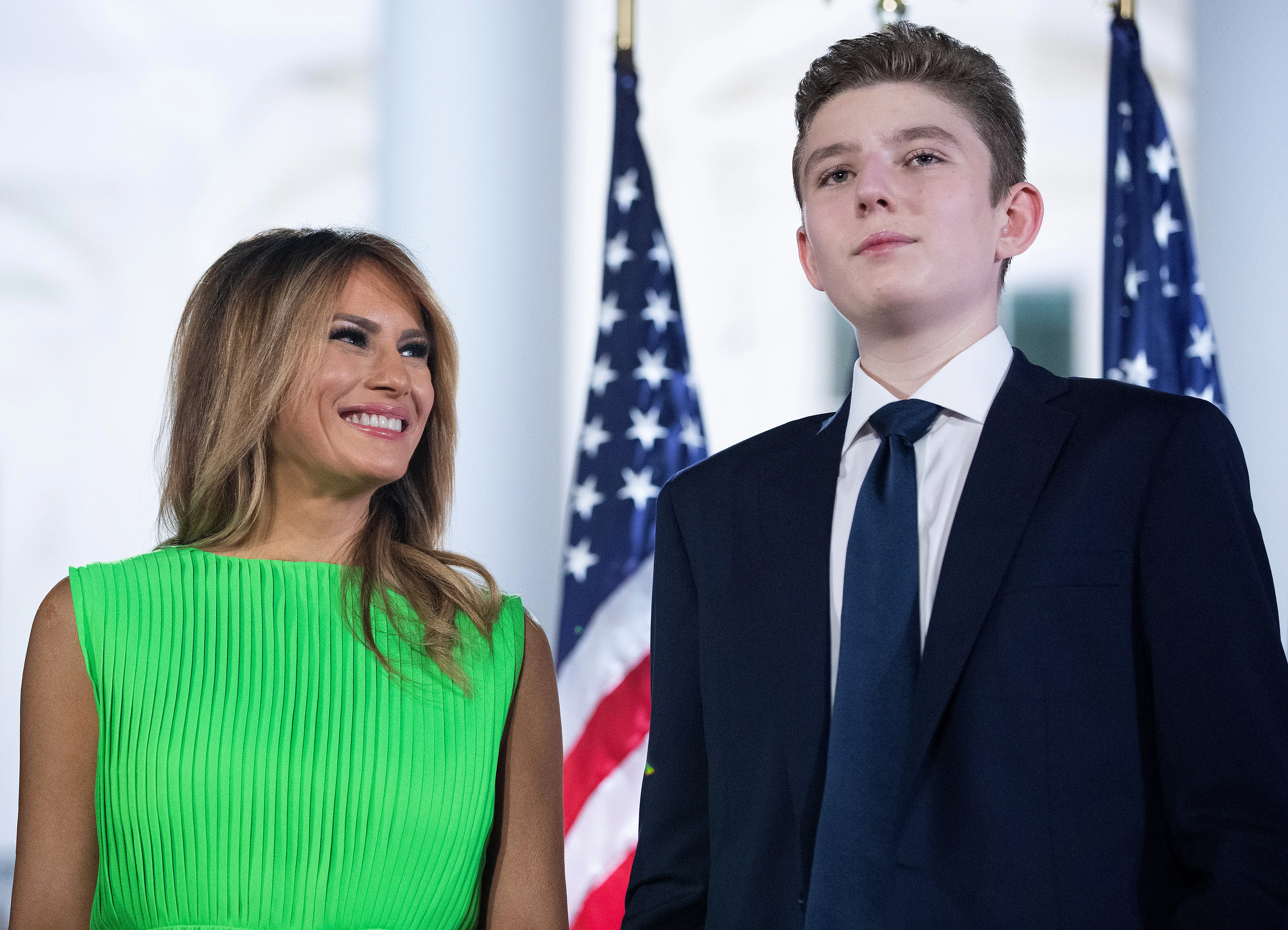 Melania Trump smiling at her son, Barron Trump, with US flags in the background.