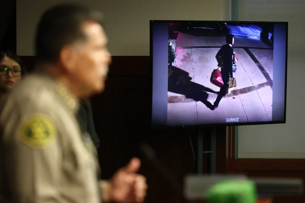 A police officer speaking while CCTV footage of a person walking on a sidewalk with a red bag and a box plays on a large screen to the right.