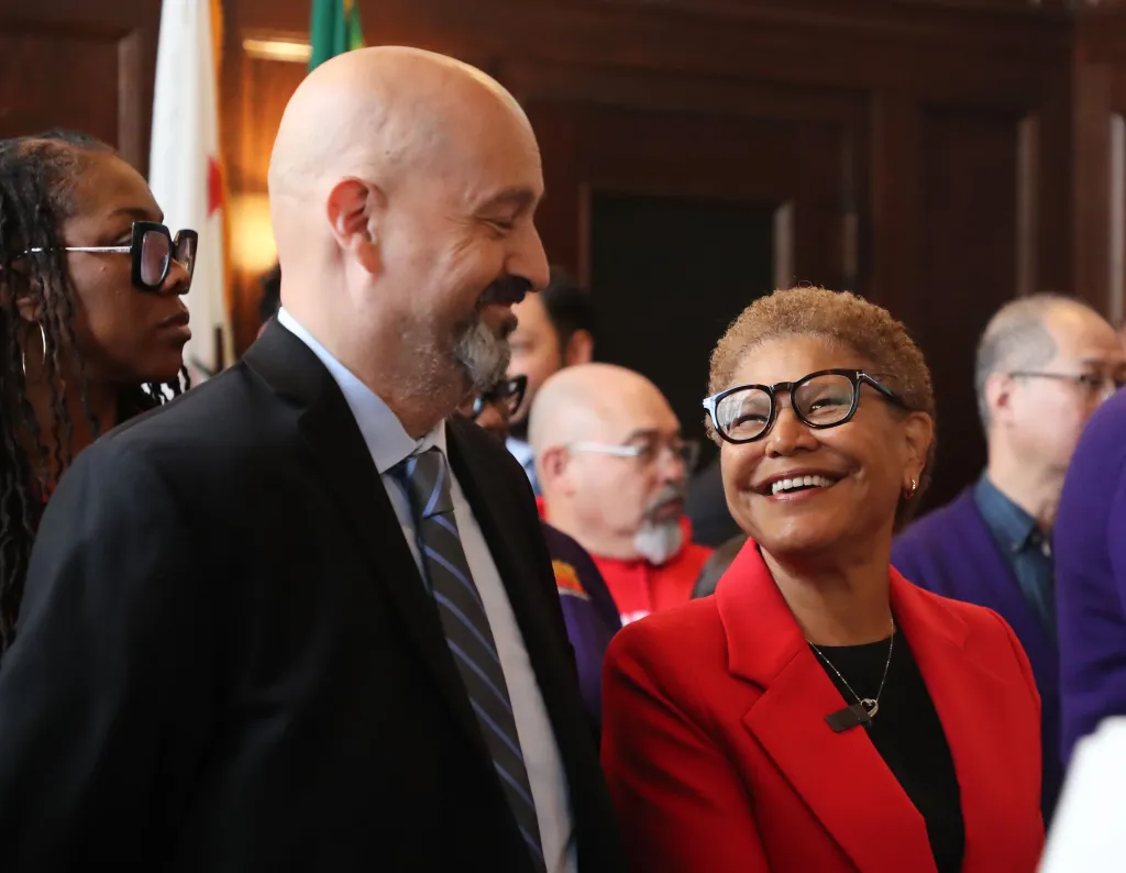 LA Mayor Karen Bass smiles and looks at the bald man standing next to her at a press conference.