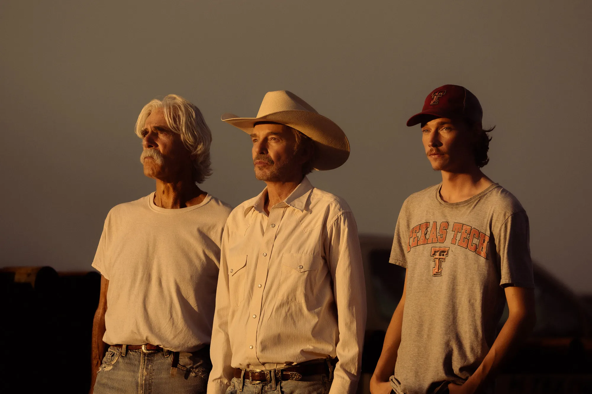 Three men, one with a cowboy hat and two with casual clothing, stand looking left.
