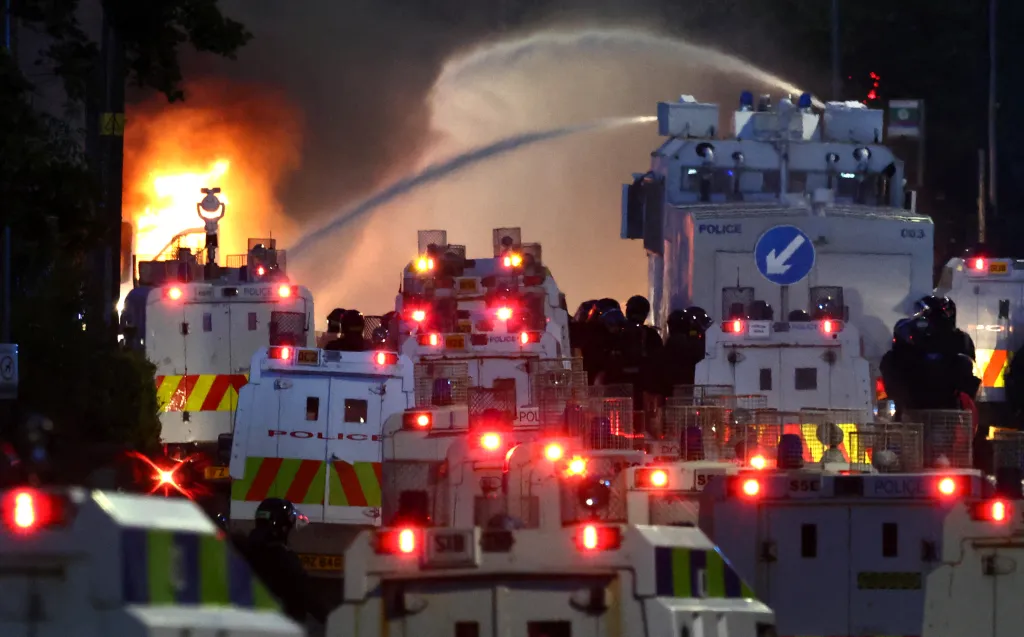 Police water cannon spraying next to a large fire and several armored police vehicles during a demonstration.