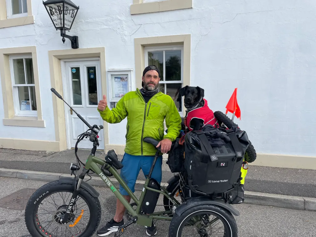 Leigh Taylor giving a thumbs up, standing next to his bike with his black dog in a red coat in a sidecar.
