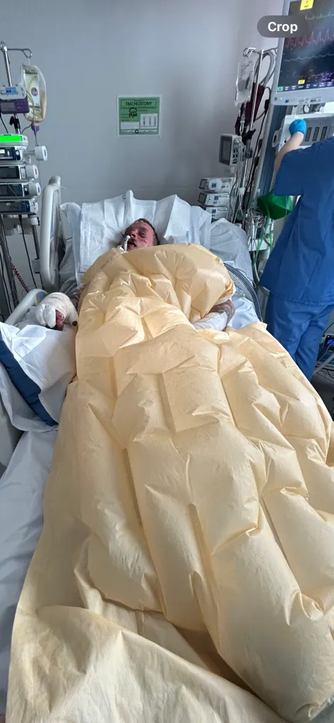 Leigh Taylor in a hospital bed with an apparent tracheostomy and bandages on his hand, covered by a light brown blanket.