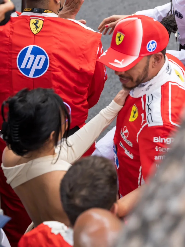 Lewis Hamilton wearing a Ferrari racing suit and cap talking to Kim Kardashian.