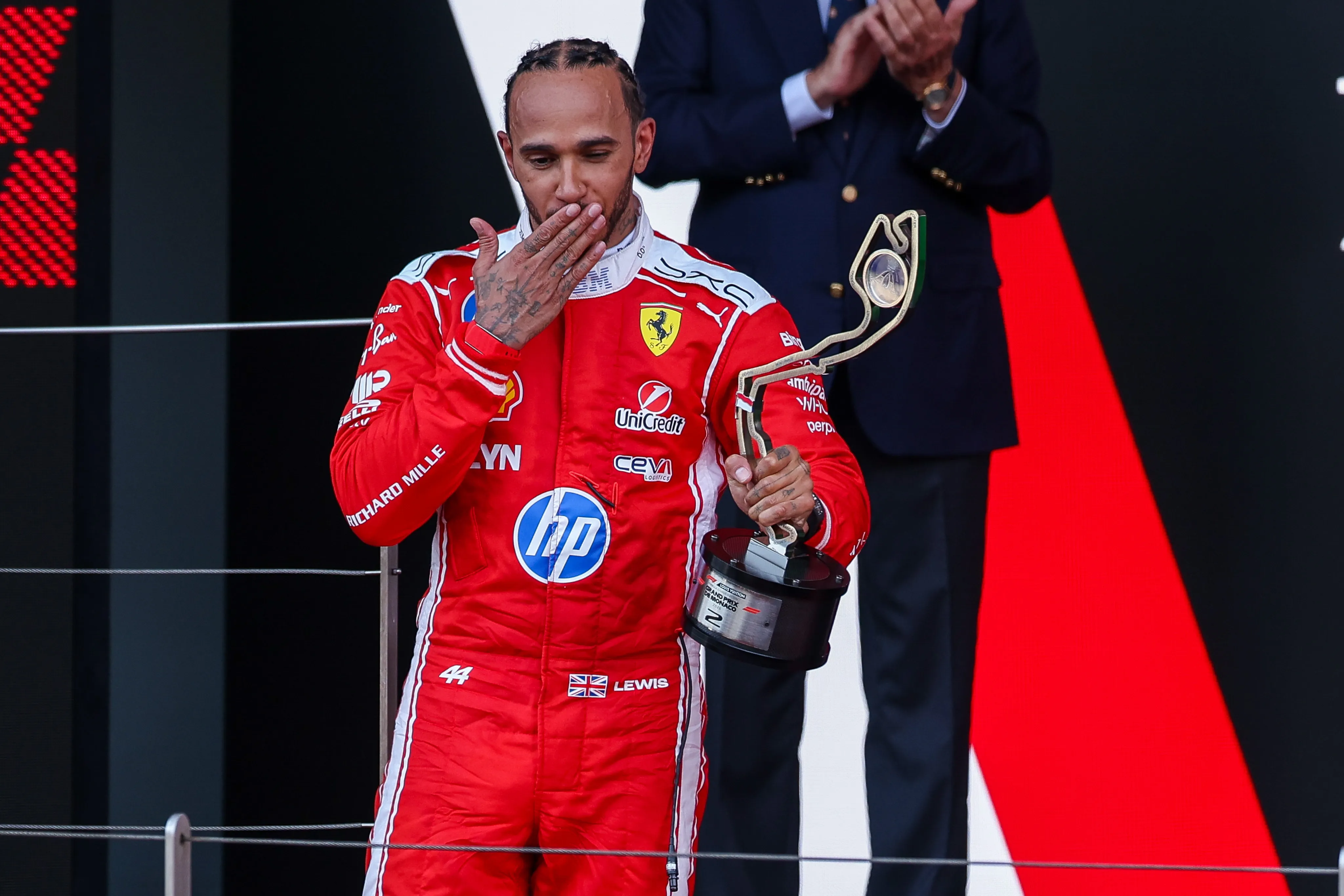 Lewis Hamilton in a red racing suit blows a kiss while holding a trophy at the Monaco Grand Prix.