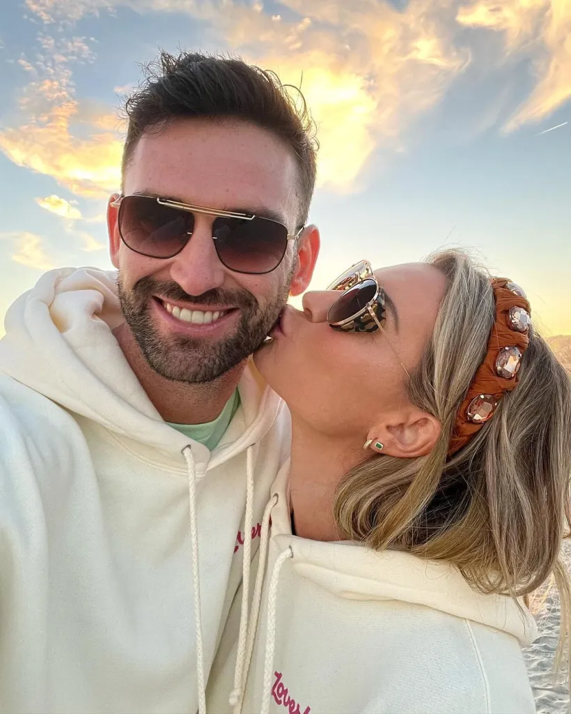 Lindsay Hubbard kissing Carl Radke on the cheek, both wearing sunglasses and white hoodies, with a sunset sky in the background.