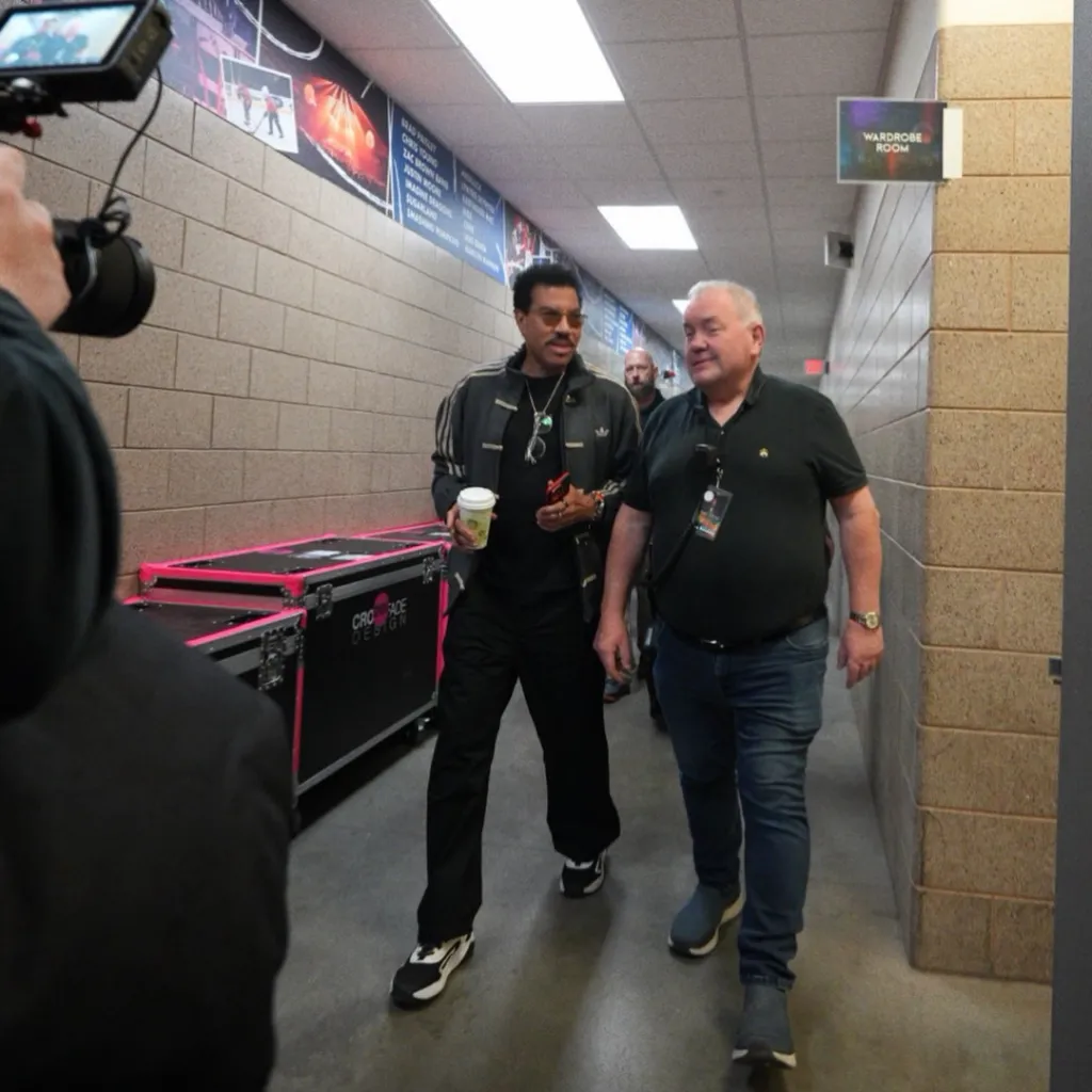 Lionel Richie walking backstage with a beverage in hand, followed by a camera crew.