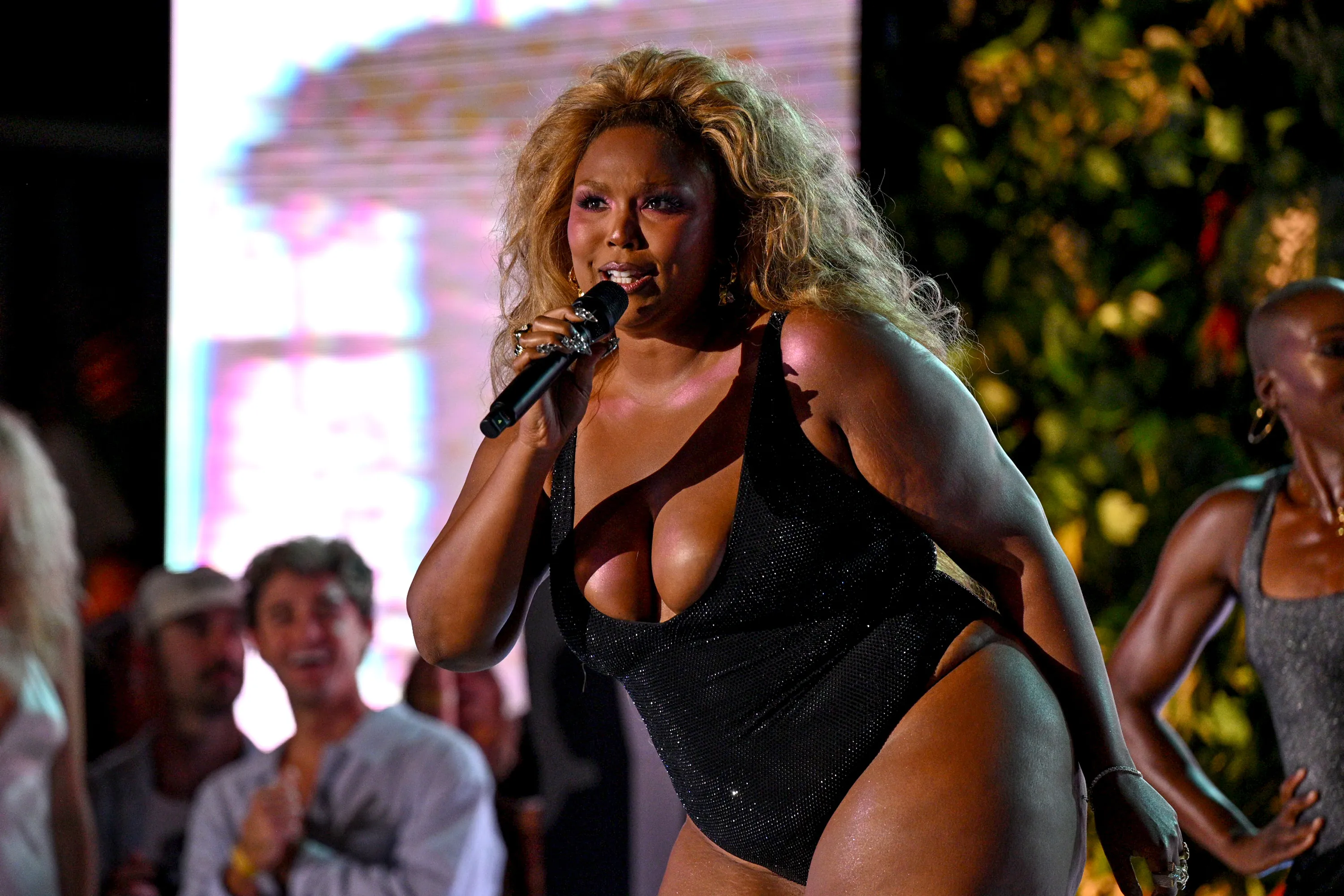 Lizzo performing on a runway in a black sequined swimsuit, holding a microphone.