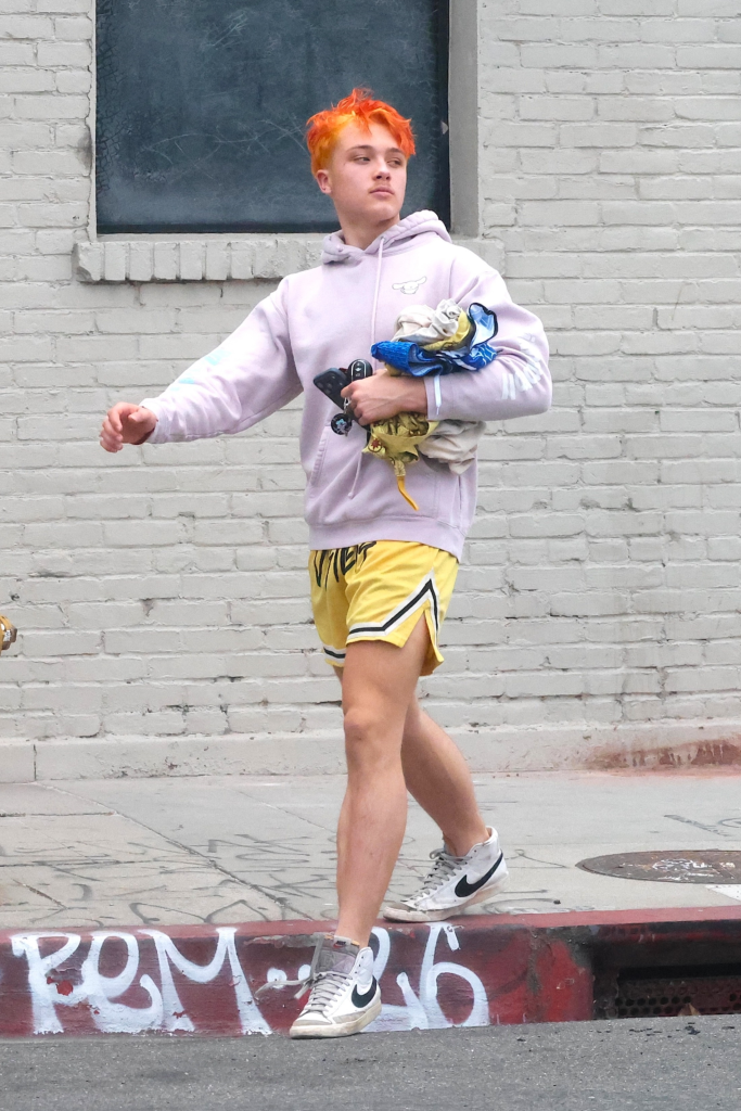 Knox Jolie-Pitt in a light purple hoodie and yellow shorts with bright orange hair, carrying clothes and a phone.