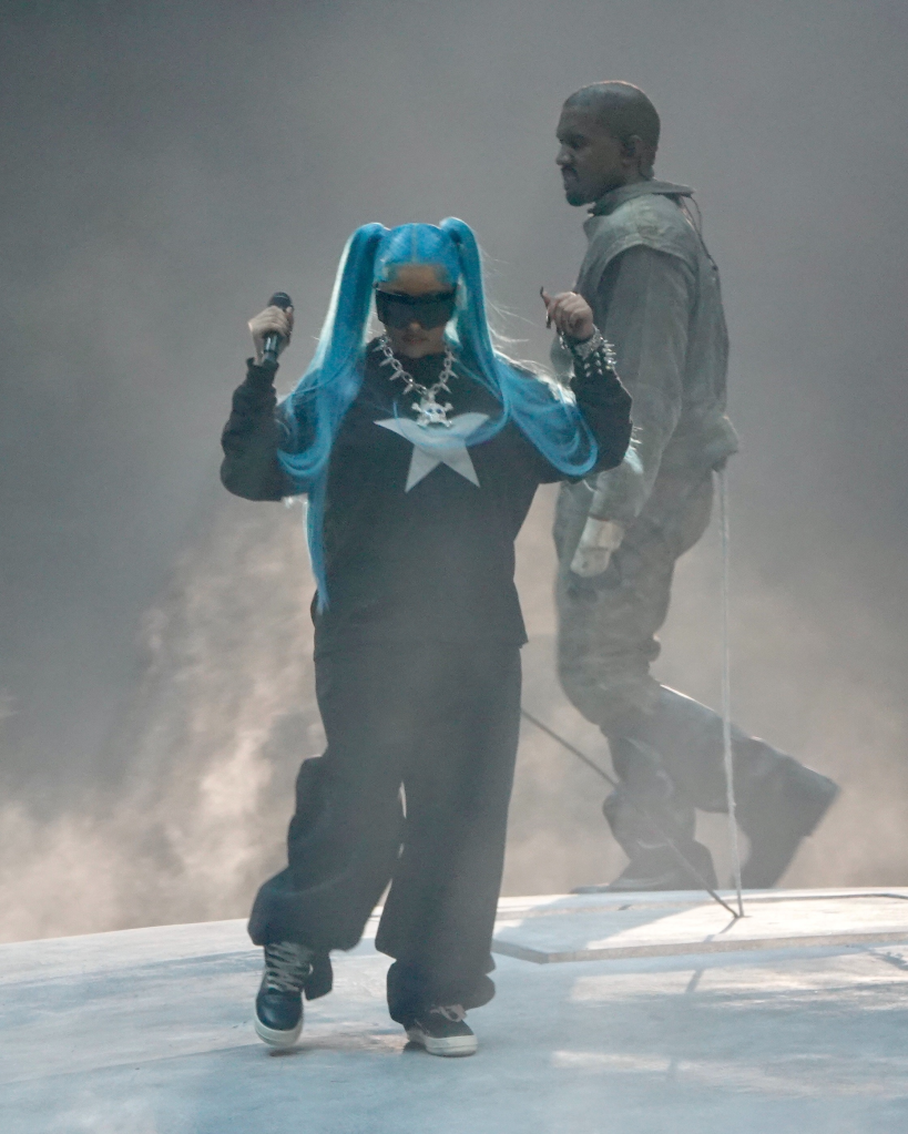 North West performing on stage with blue pigtails and a microphone, with Kanye West in the background.