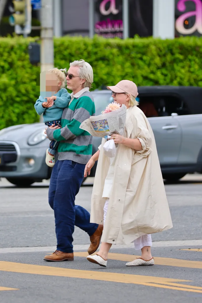Sia, her baby son, and a friend walking on a street, with Sia holding a bouquet of flowers.