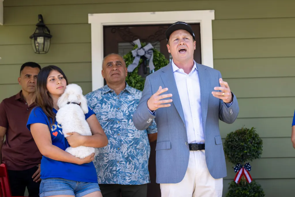 Los Angeles mayoral candidate Spencer Pratt speaks at a campaign event, with a young girl holding a white dog and two men standing behind him.