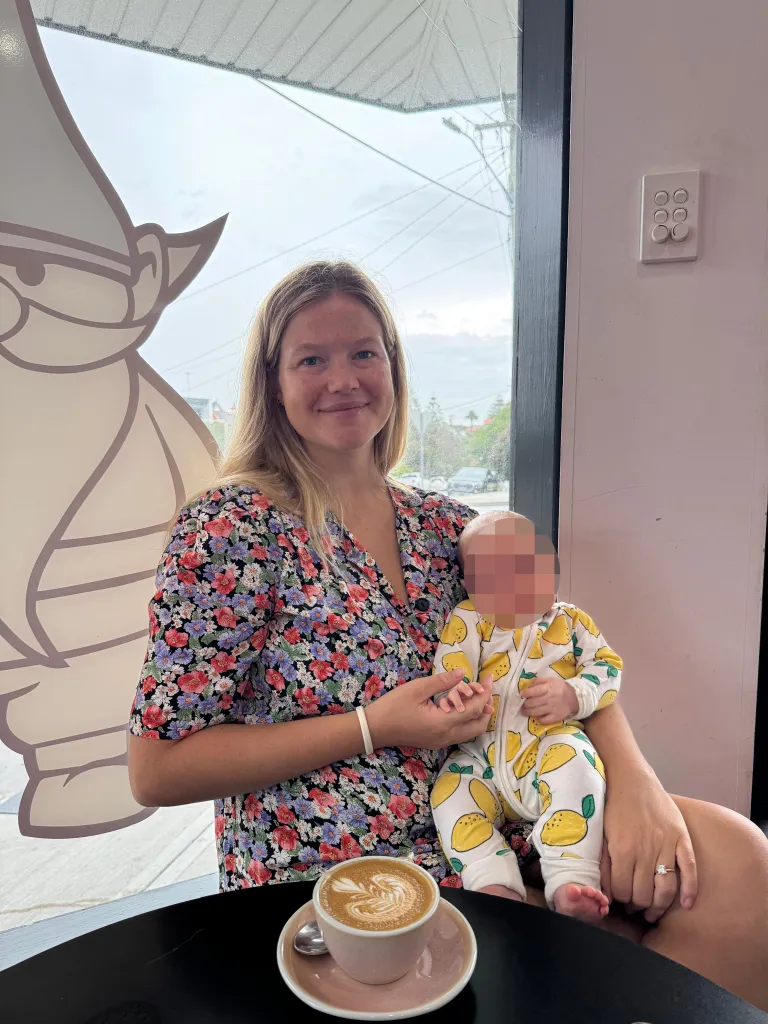 Leah Stewart holding her baby in a lemon-patterned outfit, sitting at a cafe table with a latte.