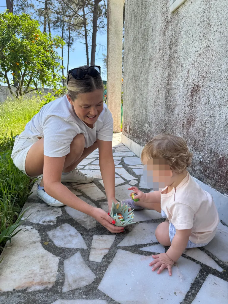 A woman crouches and holds a teal paper basket filled with colorful foil-wrapped candies for a child reaching for them.