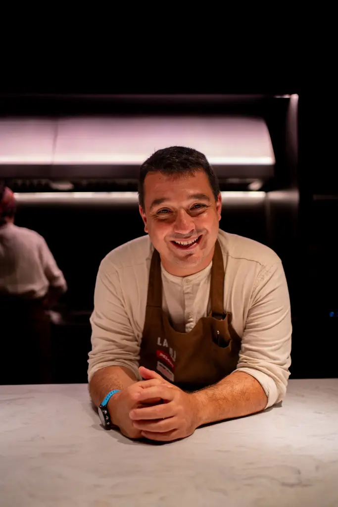 A man in a light brown apron and off-white shirt smiling at the camera.