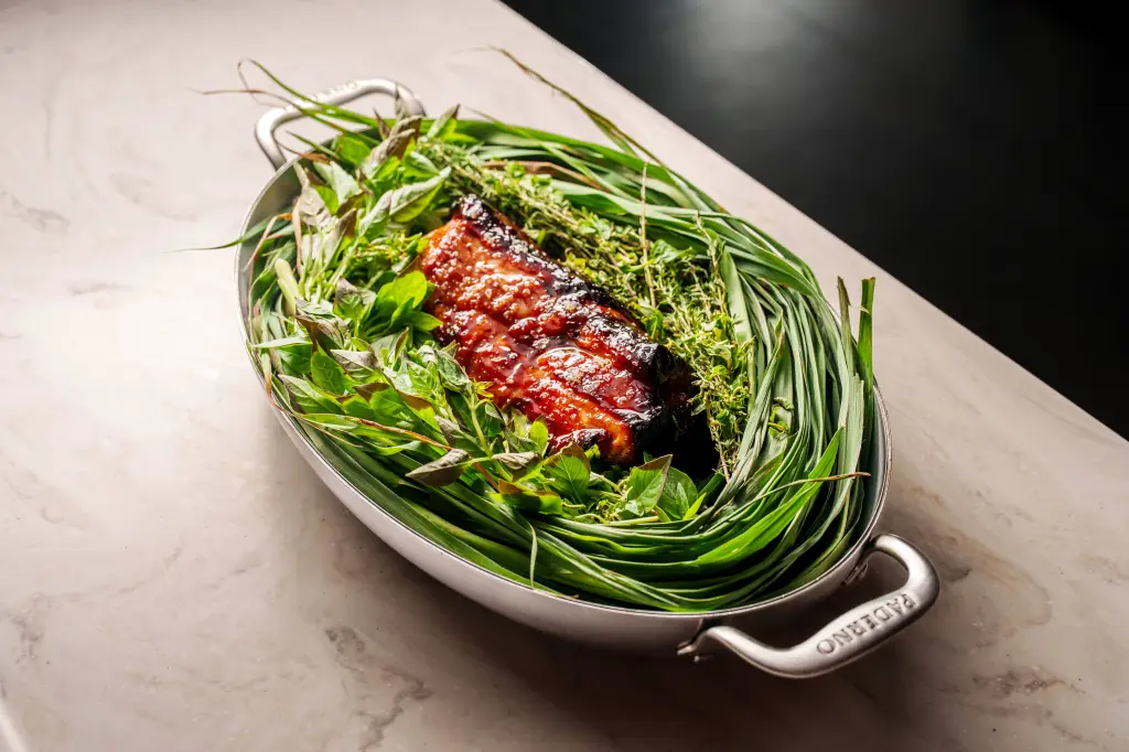 Glazed meat surrounded by fresh green herbs and chives in a silver serving dish.