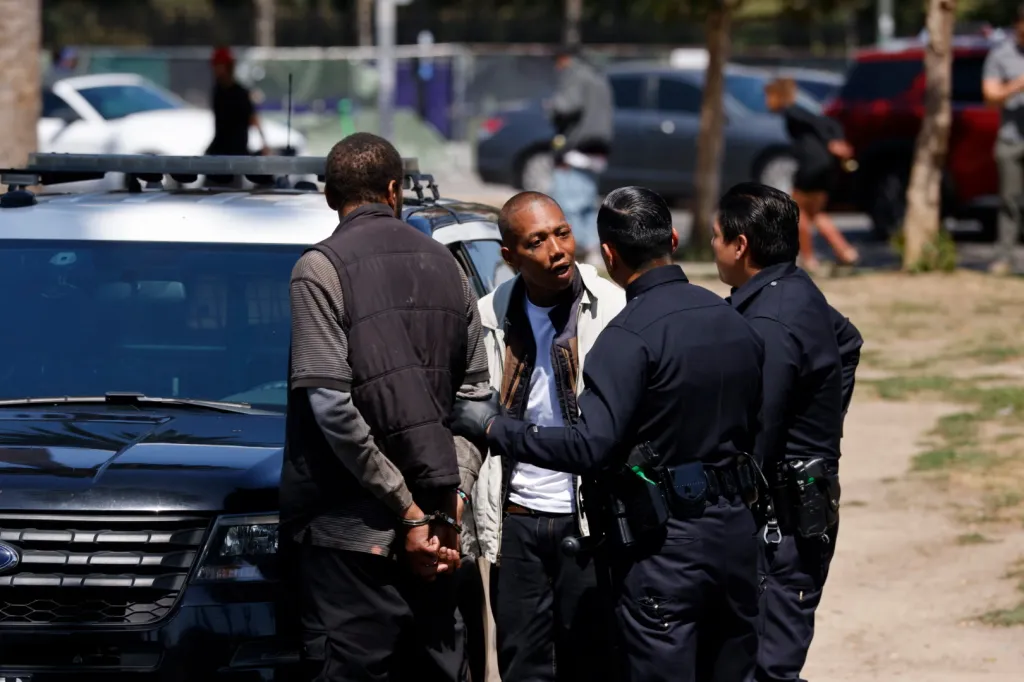 Two men in handcuffs being detained by two police officers next to a patrol car.