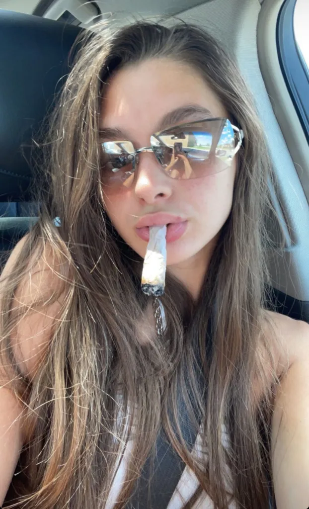Mackenzie Shirilla with long hair and sunglasses, holding a lit cigarette in her mouth.