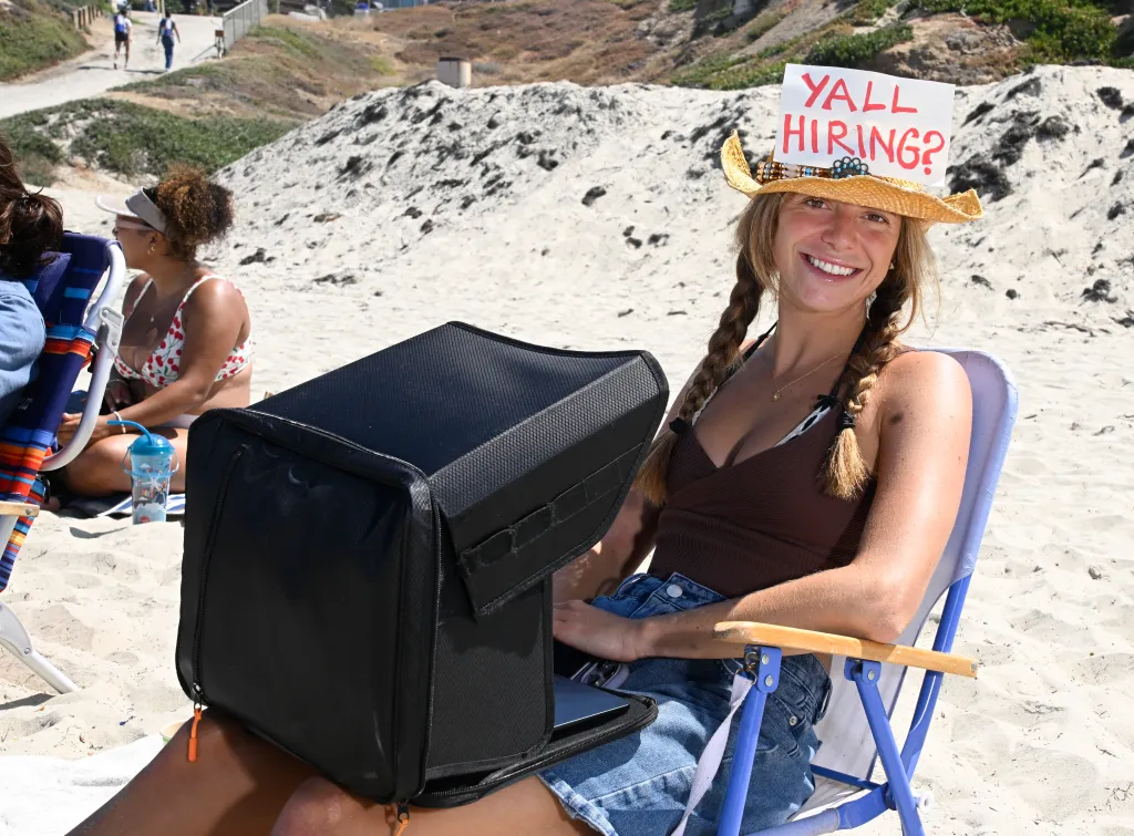 Maddy Singer works on her laptop at the beach in Pacific Beach, wearing a straw hat with a sign that says 