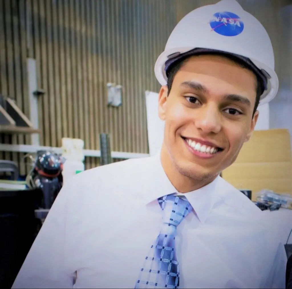 A young man in a white shirt and tie wearing a NASA hard hat smiles at the camera.