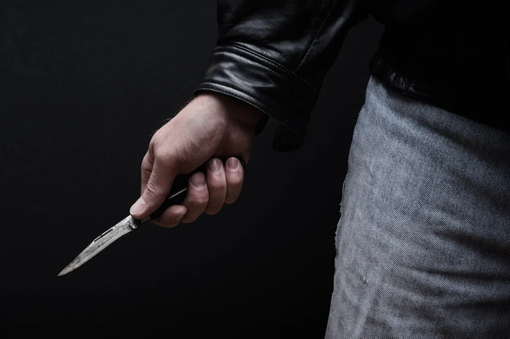 Man in a black leather jacket and jeans holding a knife with a black handle.
