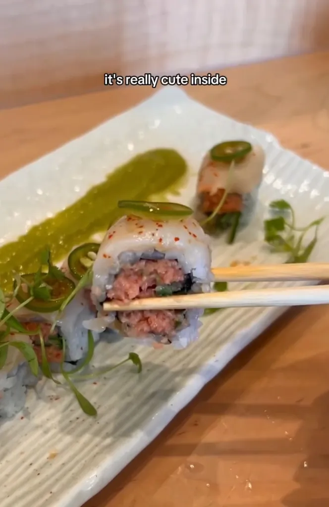 Chopsticks holding a piece of sushi roll with tuna, rice, and jalapeño on a white plate with green sauce.