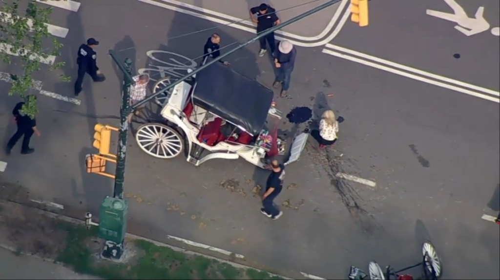 Aerial view of a horse-drawn carriage after it has flipped over in the middle of a street, with people standing around it.