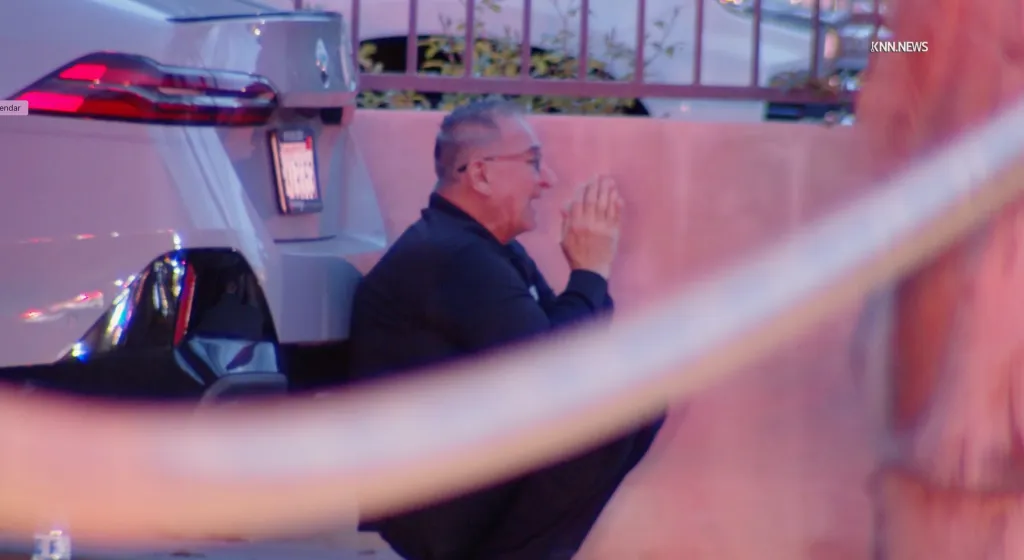 A man in glasses wearing a black shirt crying and crouching next to a white car.
