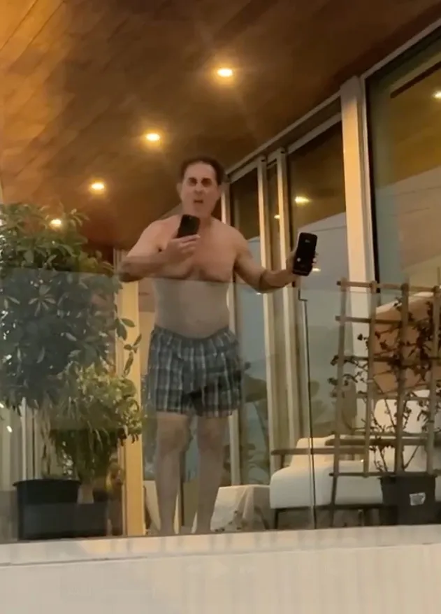 A shirtless man in boxer shorts yells racial slurs from a balcony, holding two phones.