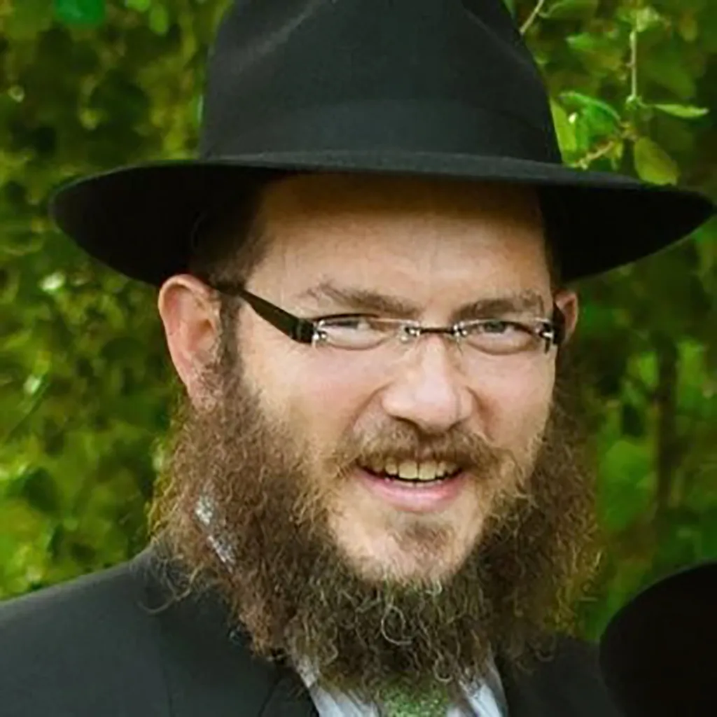 Rabbi Zushe Cunin wearing a hat and glasses, with a full beard, smiling slightly.
