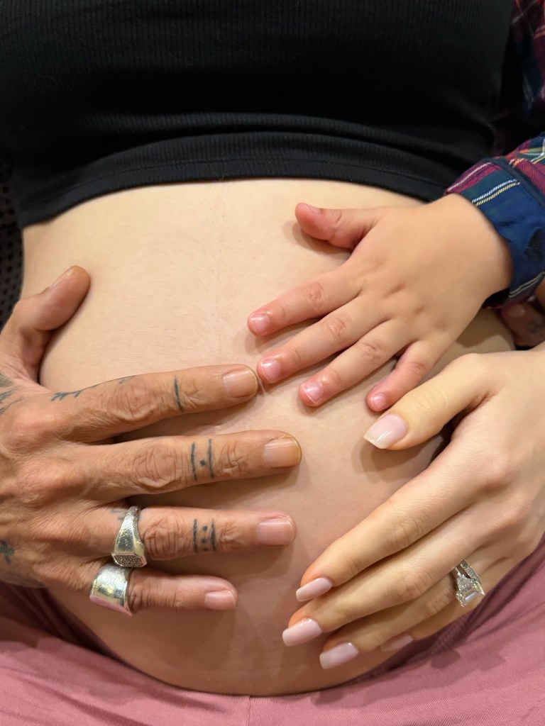 Three hands of different sizes resting on a pregnant belly.