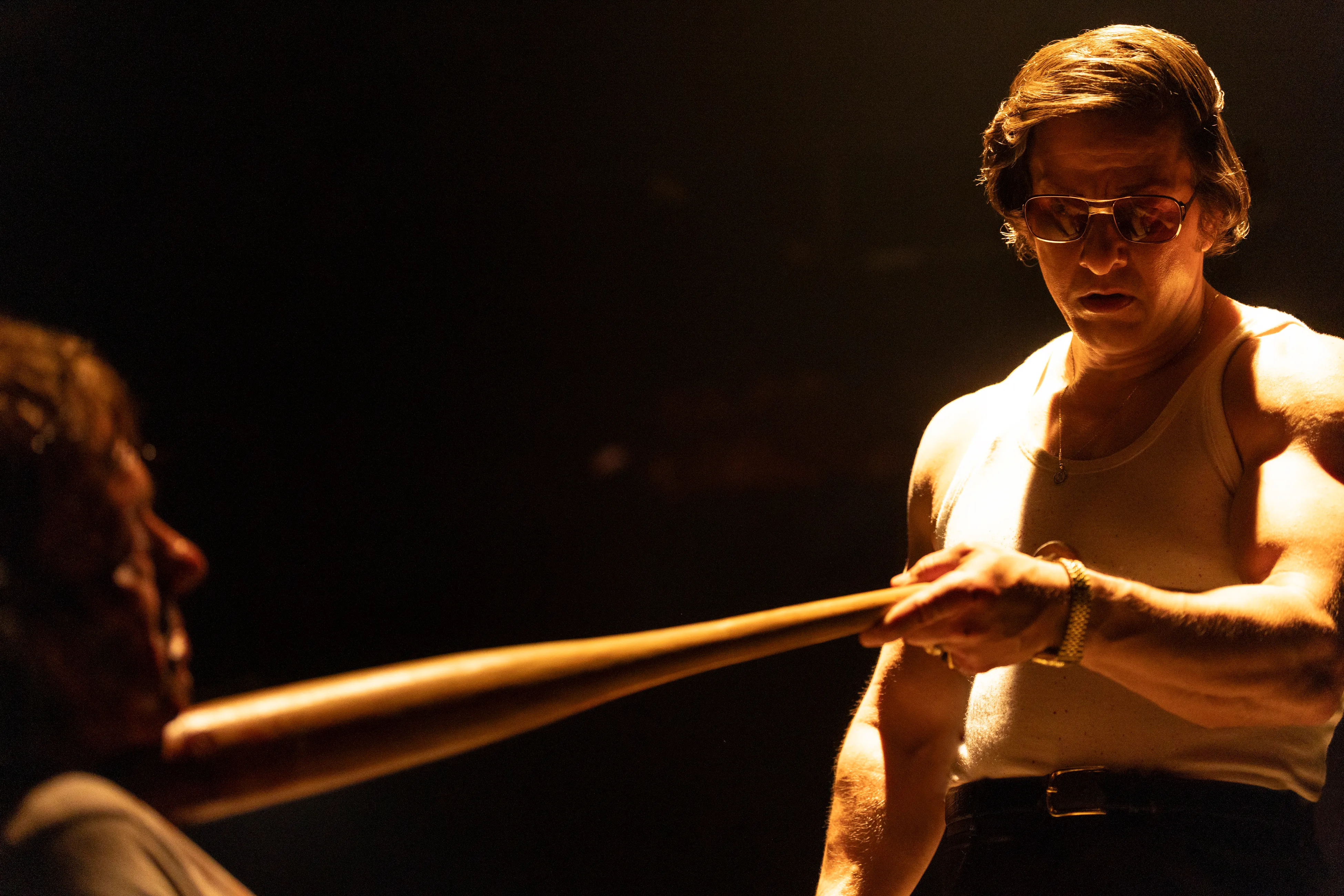 Mark Wahlberg as mafia hitman Greg Scarpa, wearing a white tank top and aviator sunglasses, holding a baseball bat.