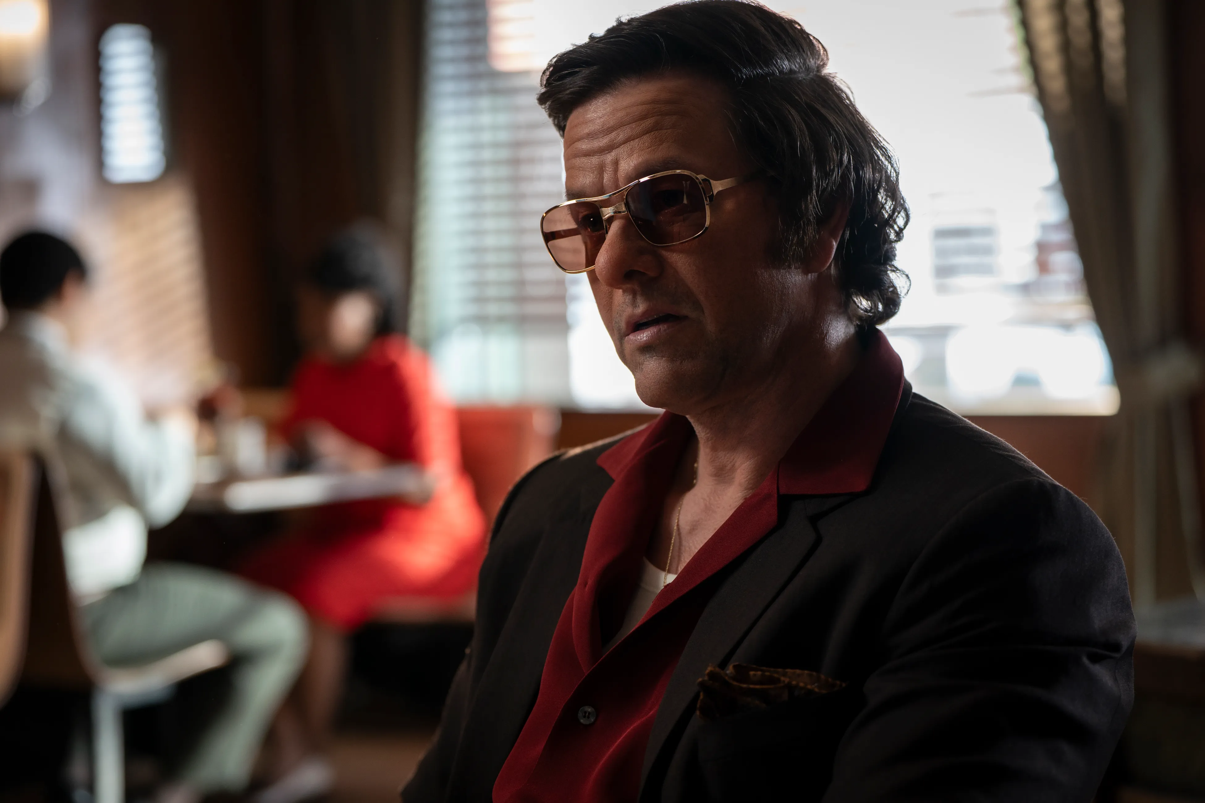 Mark Wahlberg in character as a man with dark hair, sunglasses, and a red collared shirt under a dark suit jacket.