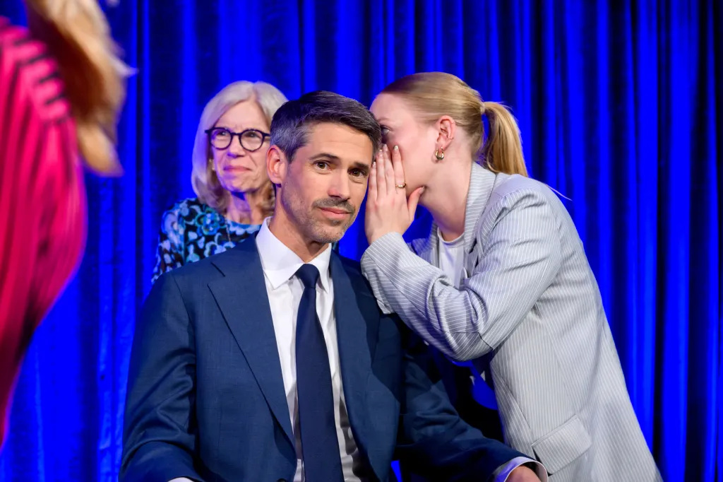 Matt Mahan, a male gubernatorial candidate, is seated on stage, looking at the camera, while a woman whispers into his ear and another woman looks on in the background.
