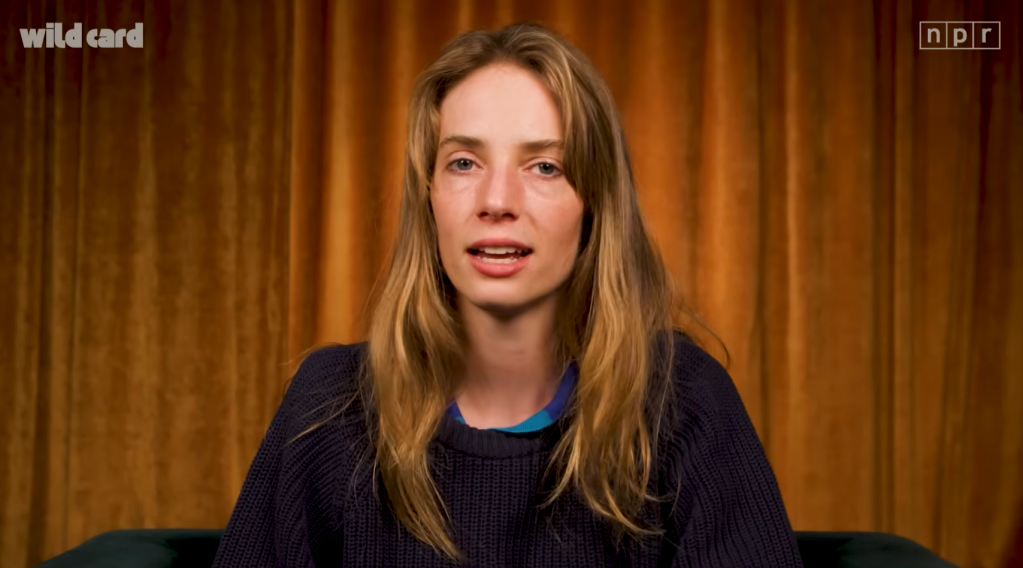 Maya Hawke, with shoulder-length blonde hair and a dark blue sweater, sitting against a golden curtain background.
