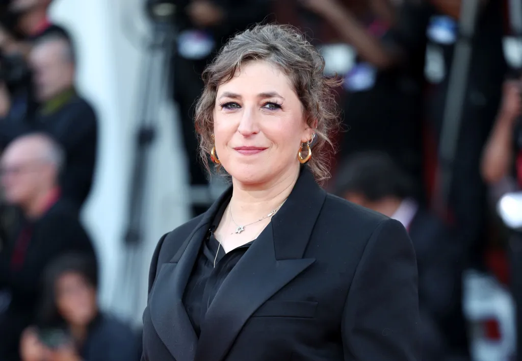 Mayim Bialik smiles while wearing a black blazer, black shirt, a necklace with a Star of David pendant, and hoop earrings.