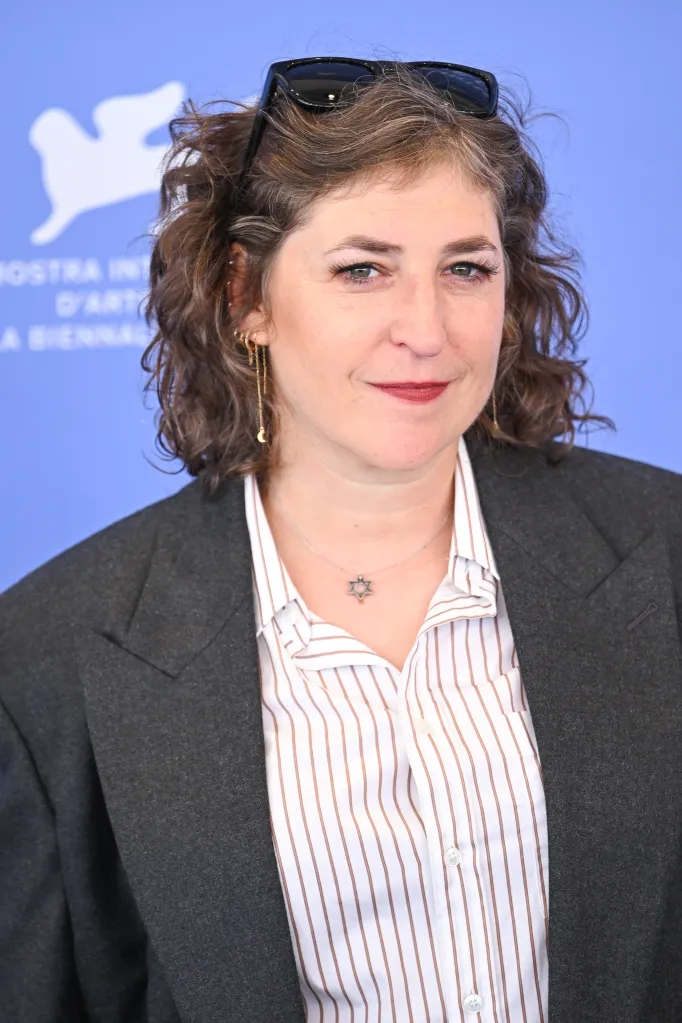 Mayim Bialik smiles, wearing sunglasses on her head, a dark blazer over a white and brown striped shirt, and a Star of David necklace.