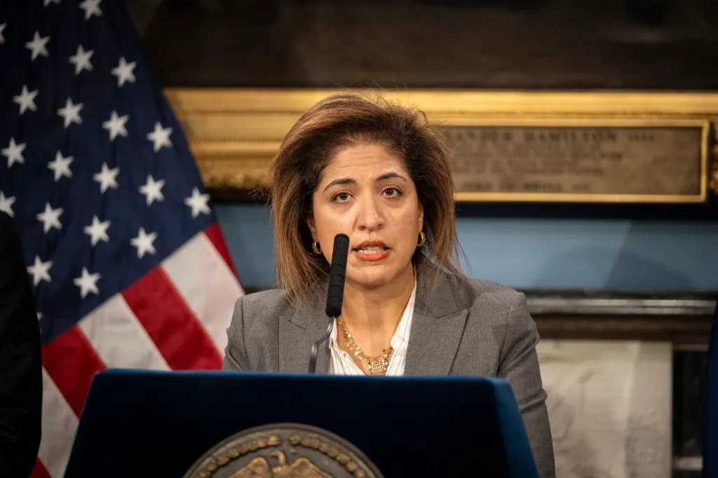 Nadia Shihata speaking at a podium with an American flag and a historical plaque in the background.
